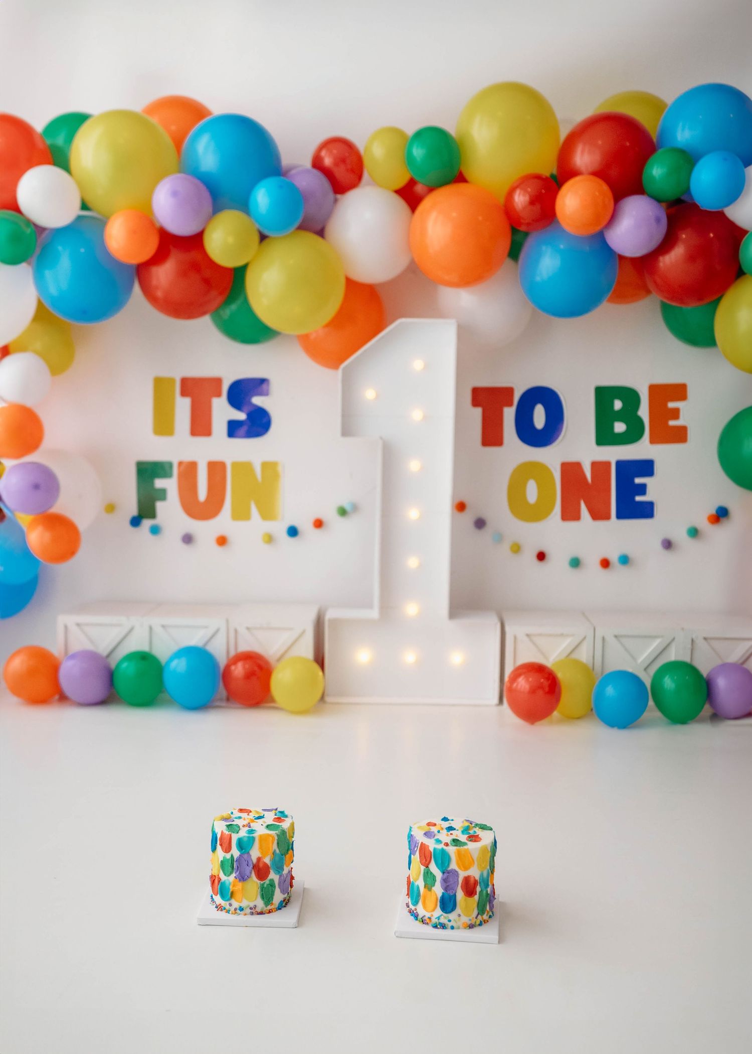 Colorful first birthday party setup with balloon garland, marquee number 1, and two decorated cakes reading Its Fun To Be One.