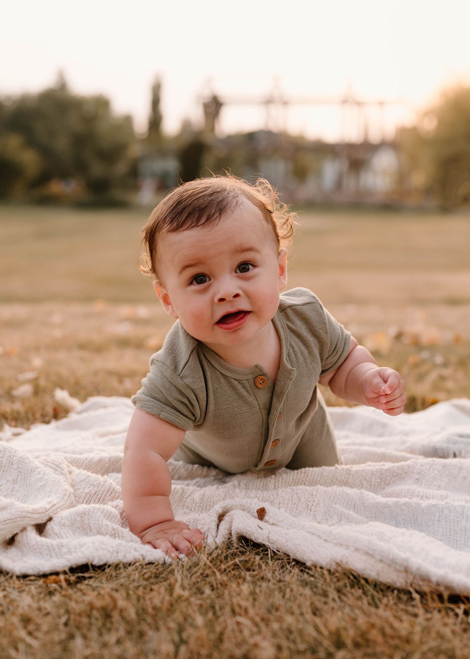 Adorable baby boy in sage green romper crawling on blanket in sunny park during golden hour.
