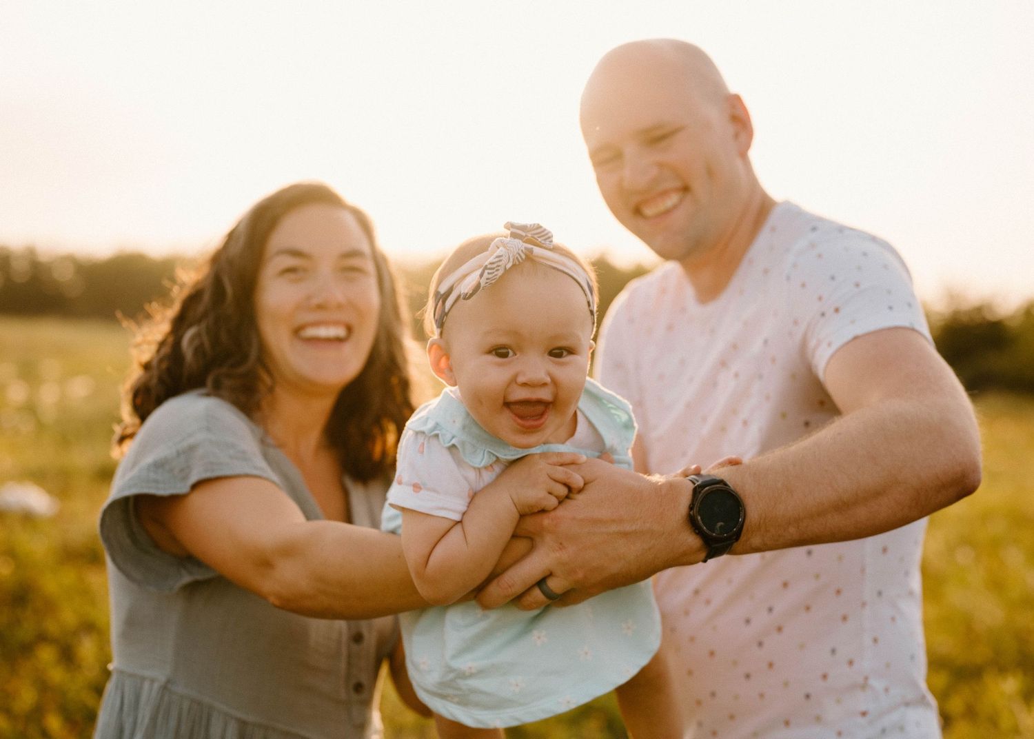 Happy young family of three smiling outdoors at golden hour, parents holding their cheerful baby girl.