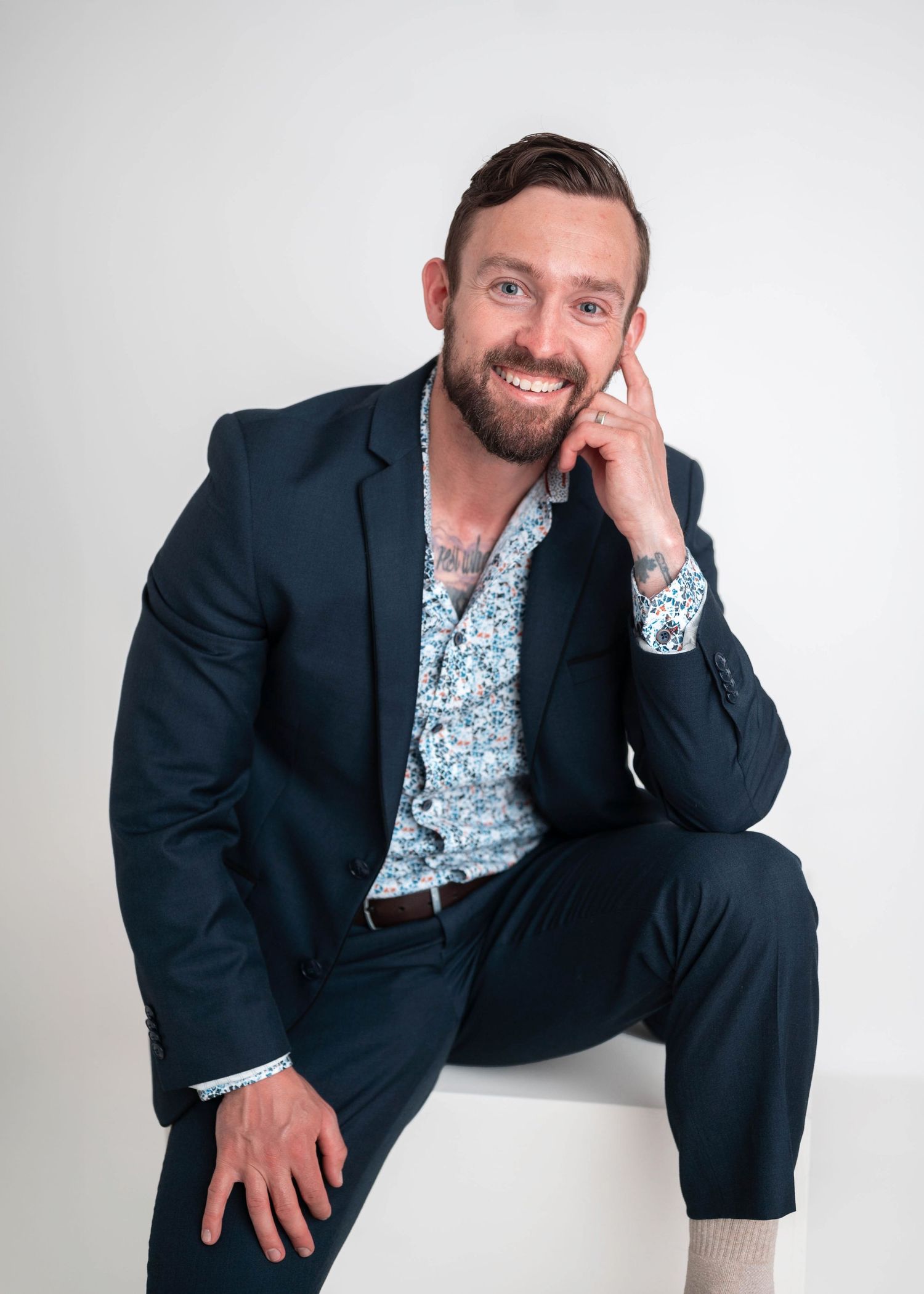 Man in navy suit and floral shirt sits smiling confidently against white background for professional headshot.