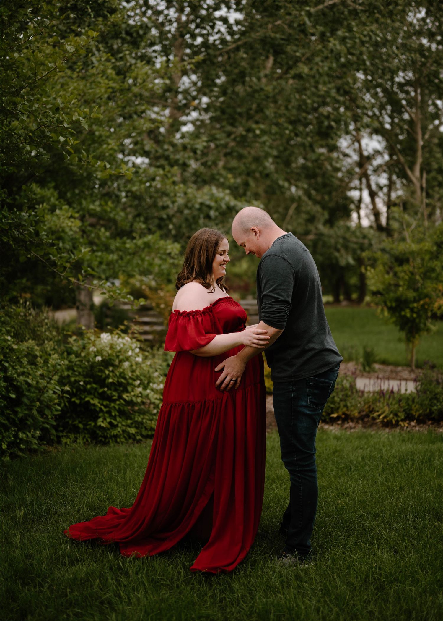 Couple in garden, woman in red maternity gown, man in dark shirt, sharing tender moment on green lawn.