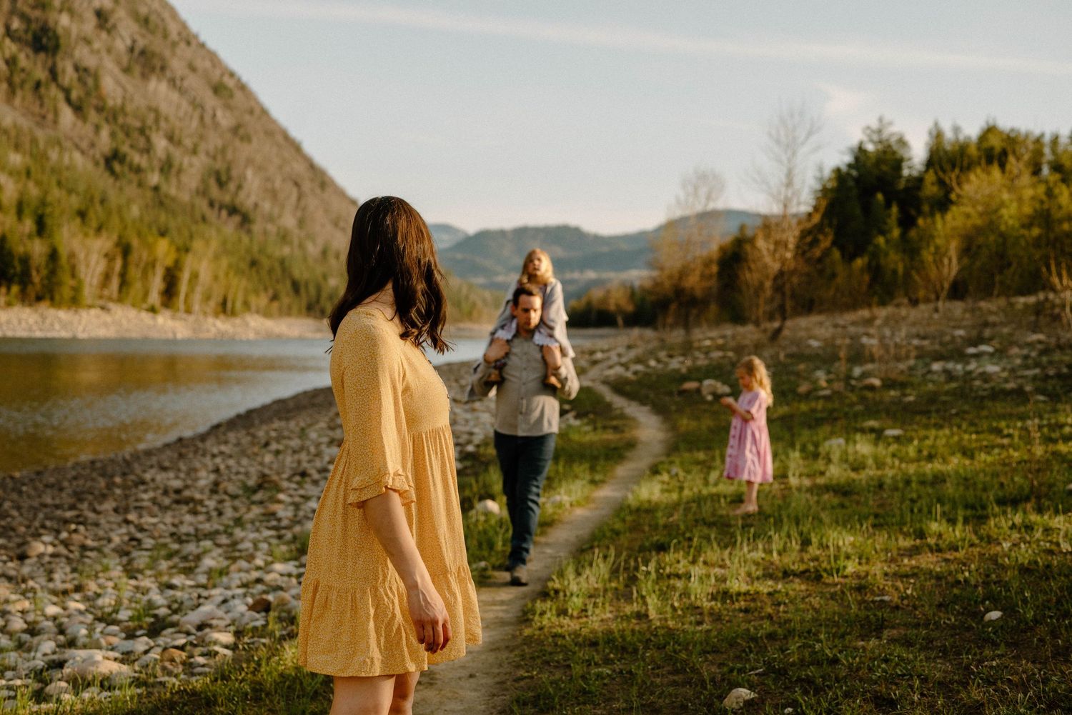 Family enjoying a peaceful walk along a rocky riverbank with mountains in the background.