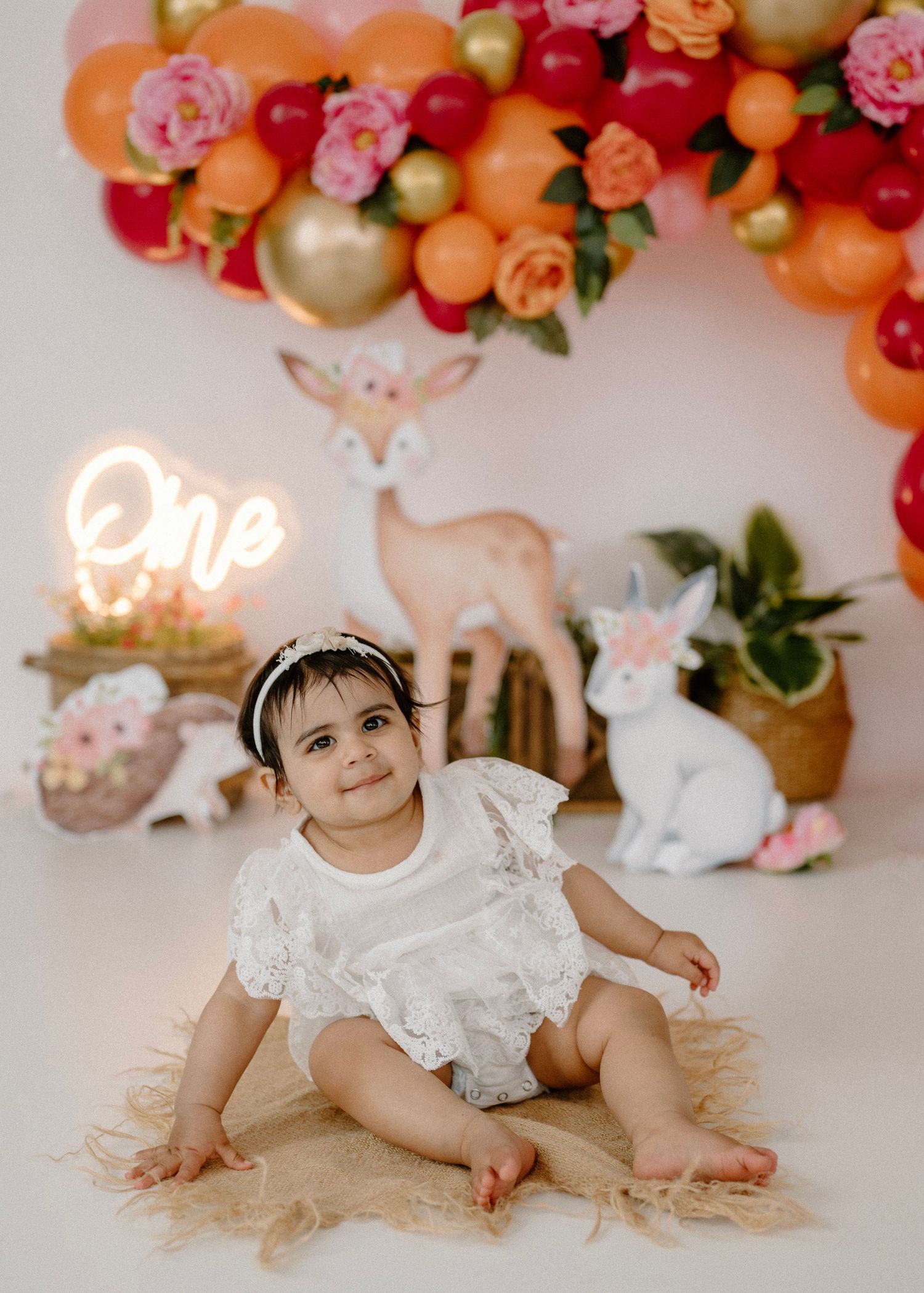 Smiling baby girl in white romper sitting at first birthday party with colorful balloon arch.