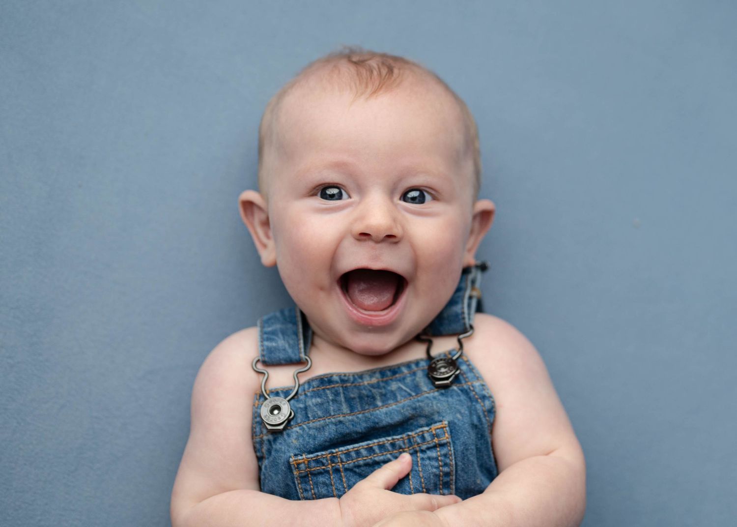 Laughing baby in denim overalls lying on a blue background with a joyful expression.