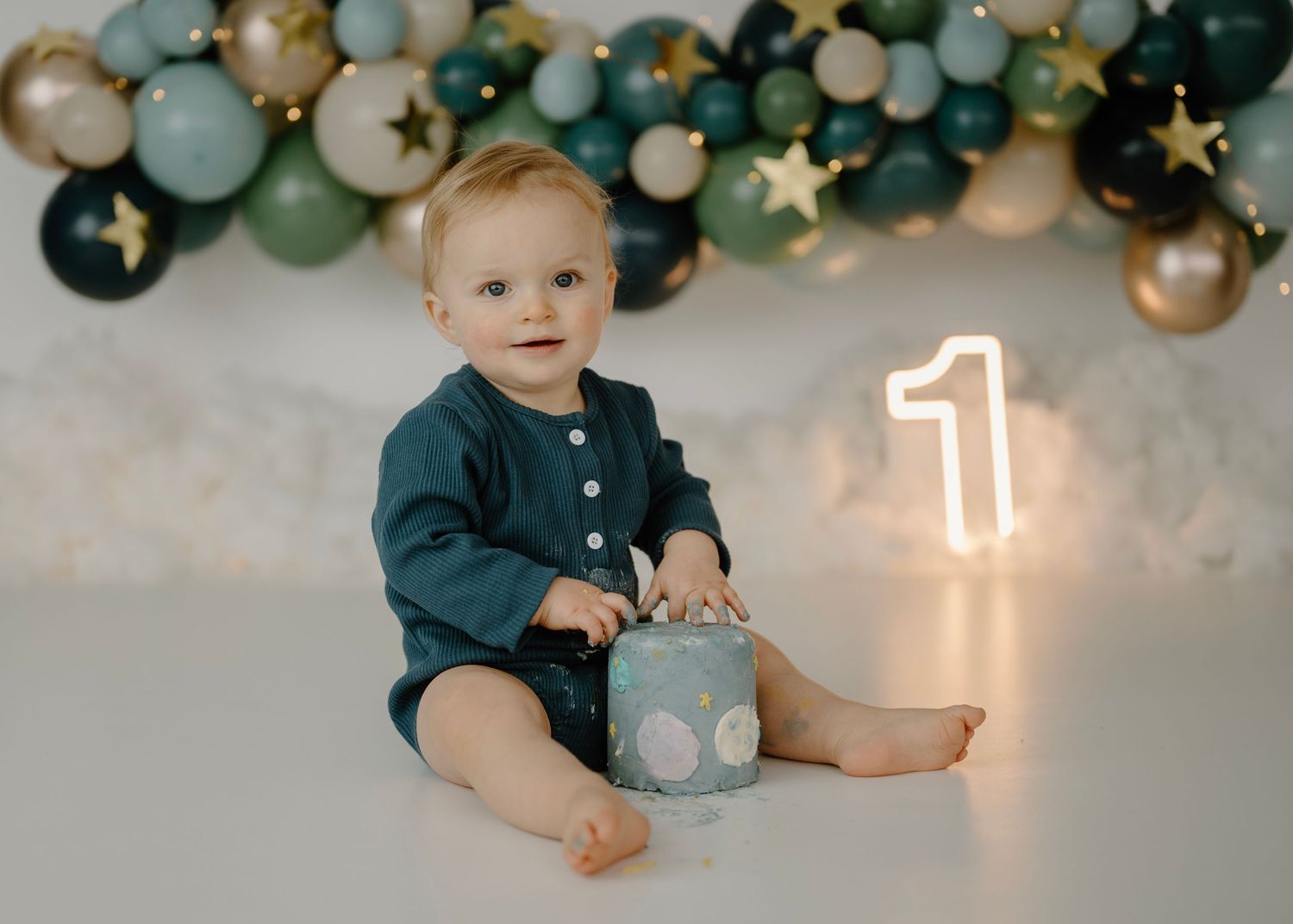 Baby boy in teal romper sitting with small cake beside glowing number 1 and balloon garland.