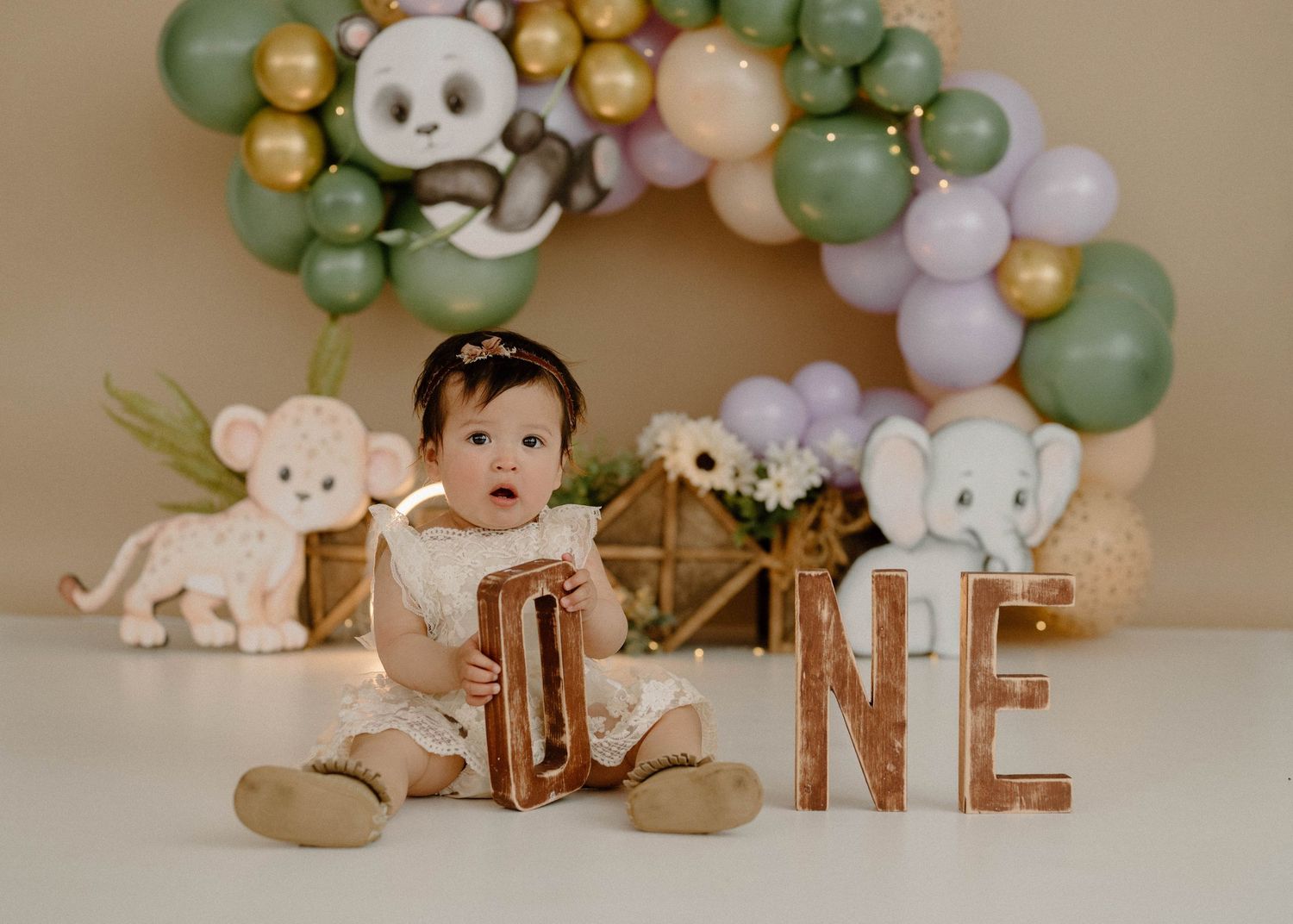 Baby girl in white dress holds ONE letters with safari animal balloon arch backdrop.