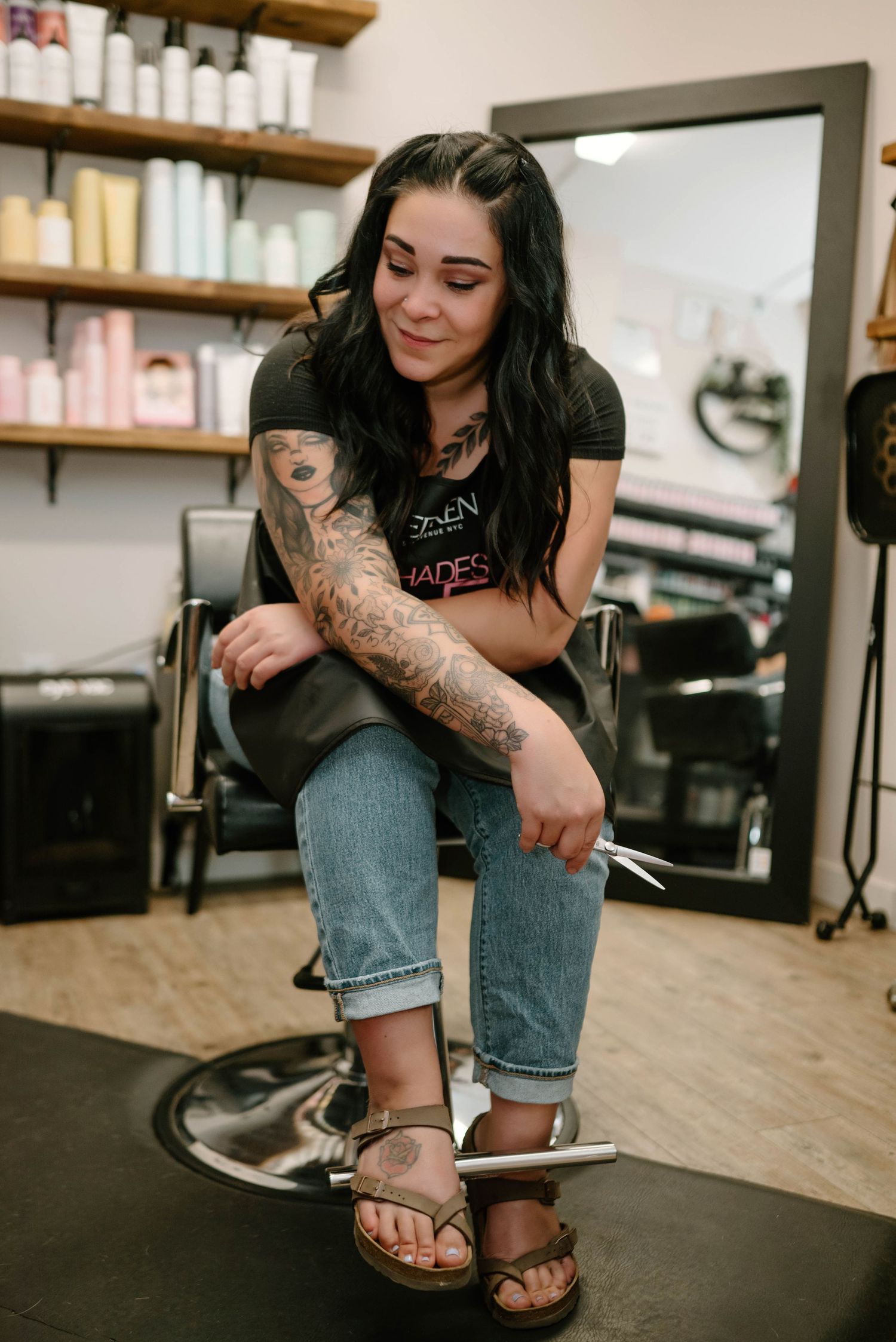 Tattooed female hairstylist sitting in salon chair holding scissors in hair salon.