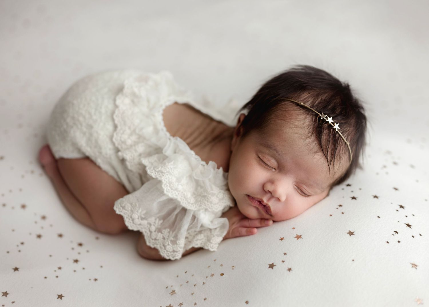 Sleeping newborn baby in white lace romper and floral headband rests peacefully on a soft white backdrop.