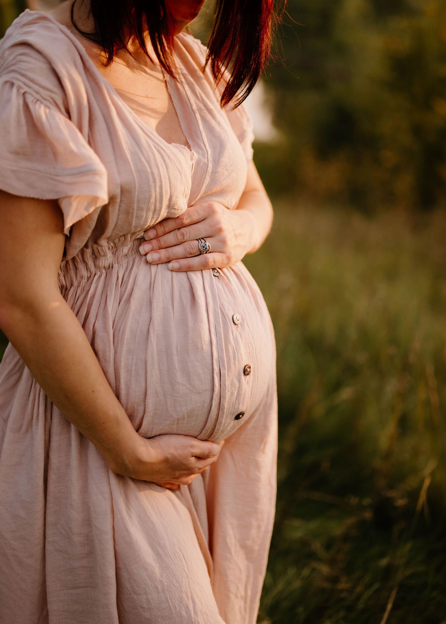 Close-up of pregnant woman in blush dress gently holding her baby bump outdoors.