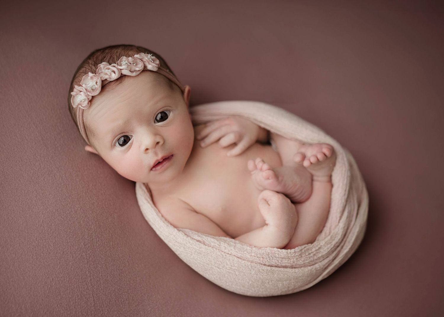 Newborn baby wrapped in soft pink fabric with floral headband, posed on mauve background in professional portrait.