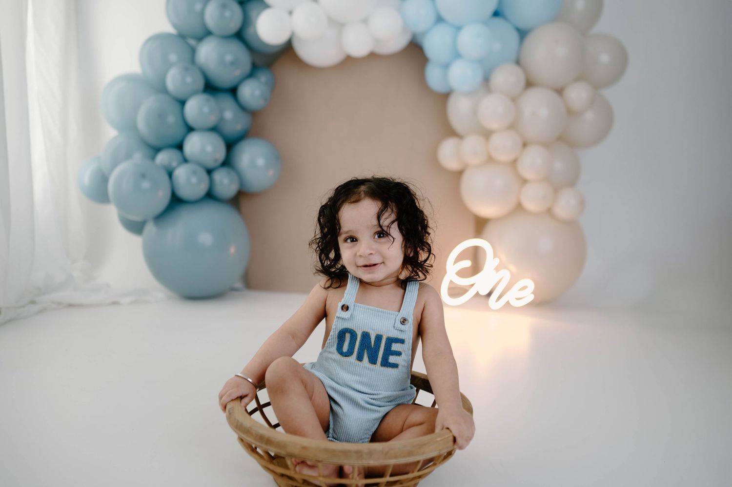 Happy baby in blue ONE romper sits in wicker basket with blue and white balloon arch behind them.