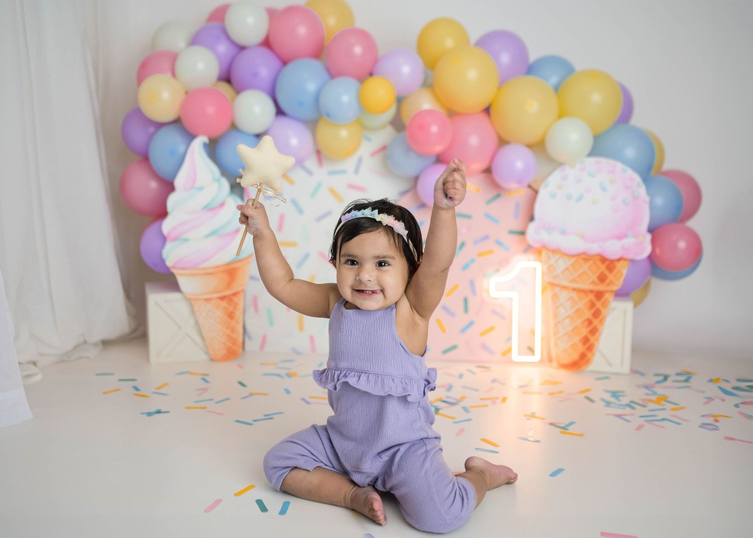 Joyful baby in lavender romper raises arms at ice cream themed first birthday with pastel balloon garland.