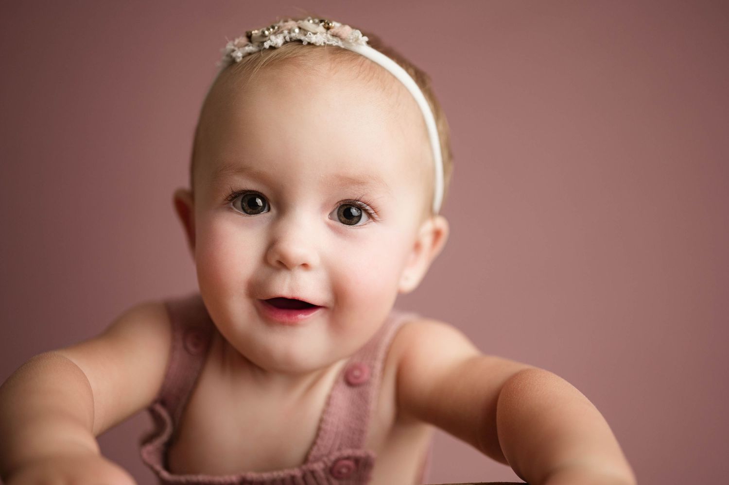 Adorable baby girl in pink romper and jeweled headband smiles during professional studio photography session.