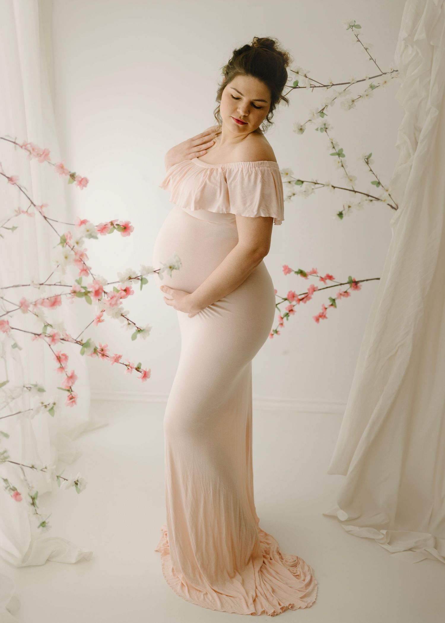 Elegant pregnant woman in blush form-fitting off-shoulder gown poses among pink cherry blossom branches in studio.