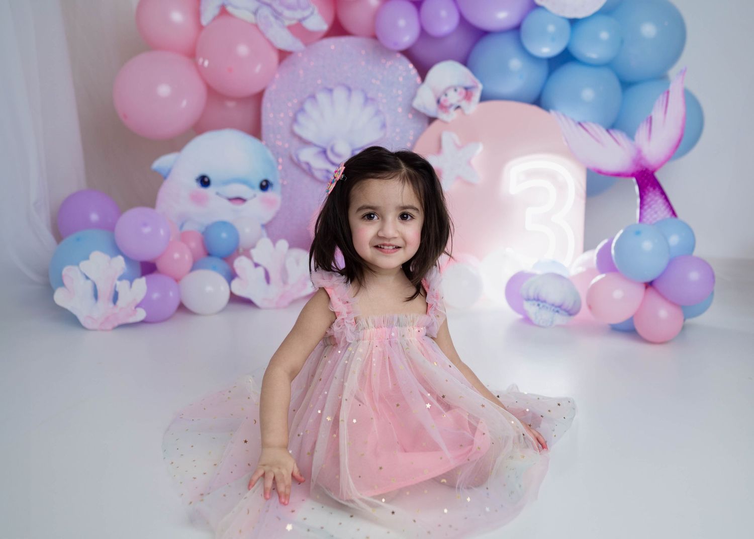 Smiling toddler girl in pink tulle dress sitting before mermaid themed balloon birthday backdrop.