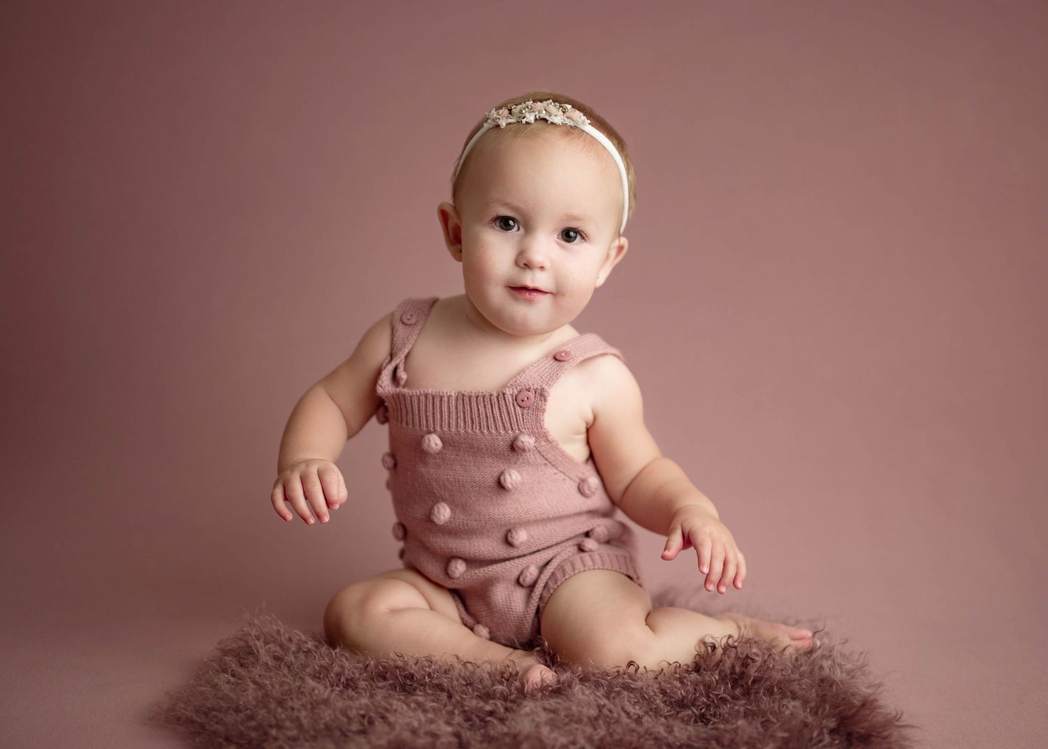 Baby girl in a mauve knit romper sitting on a fur rug against a dusty rose studio backdrop.
