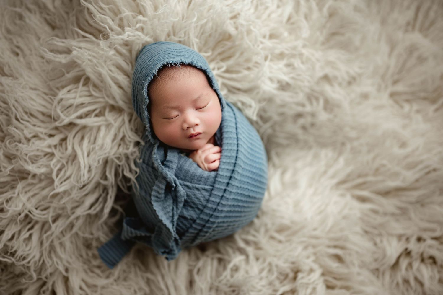 Sleeping newborn baby swaddled in blue knit wrap with matching bonnet resting on a fluffy beige flokati rug.
