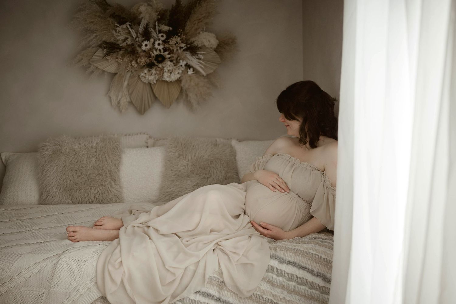Serene pregnant woman in cream dress lying on bed cradling her baby bump in a softly decorated room.