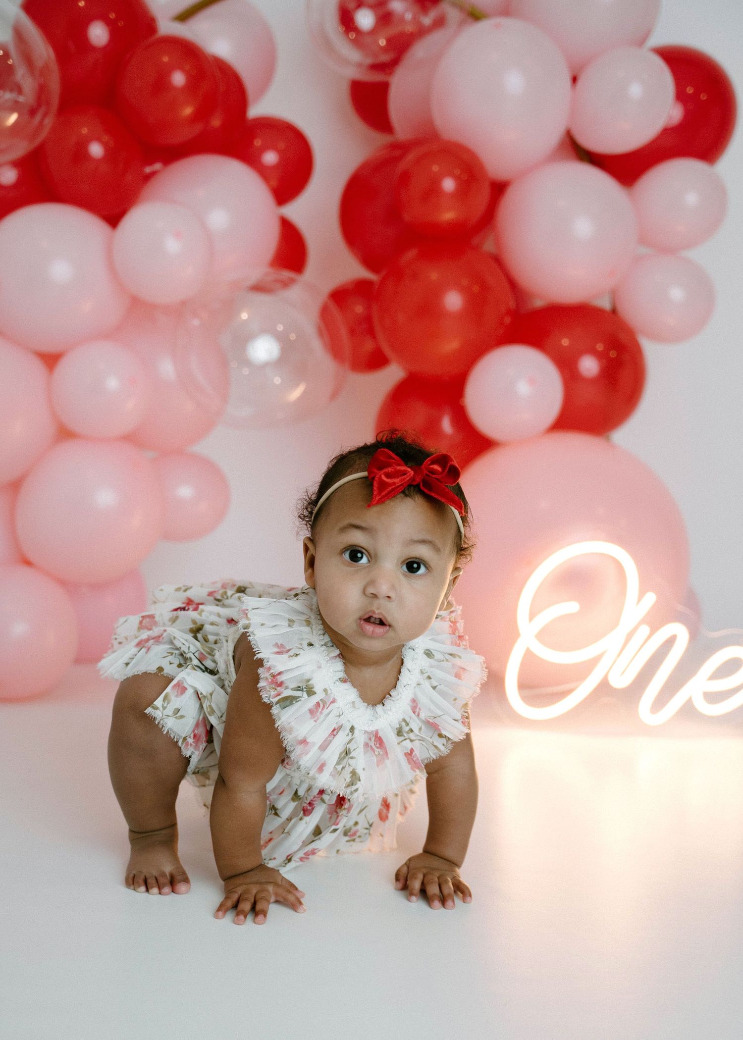 Baby girl in floral romper crawls near pink and red balloon garland with glowing One neon sign.