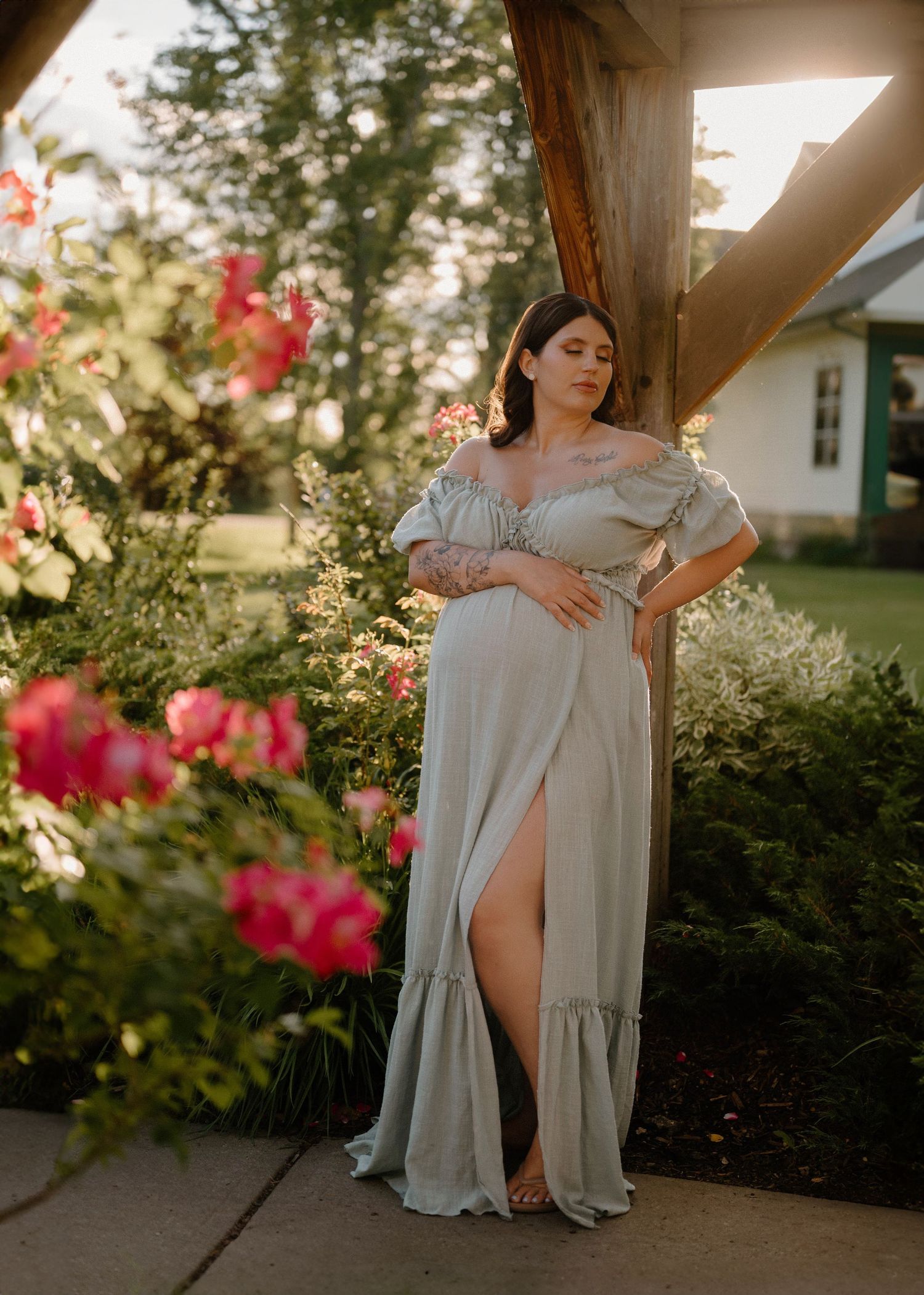 Pregnant woman in sage green off-shoulder maxi dress poses gracefully among blooming roses in golden hour light.