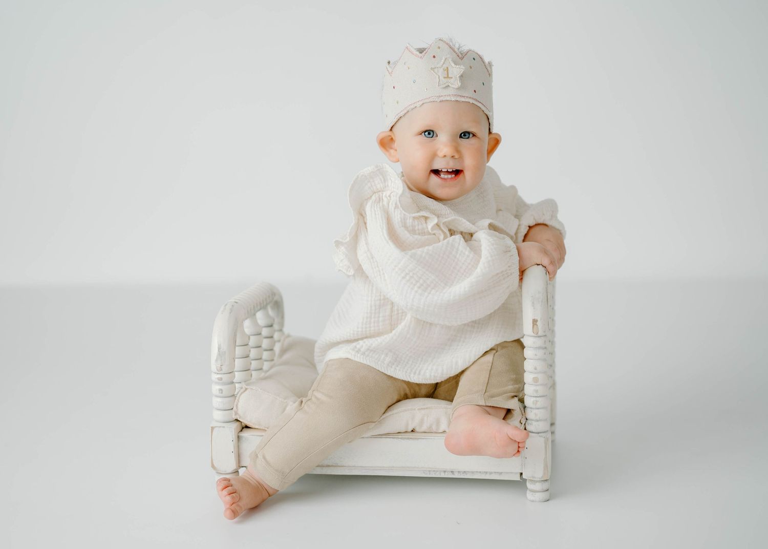 Cheerful baby wearing cream crown and white outfit sitting on small white chair in studio.