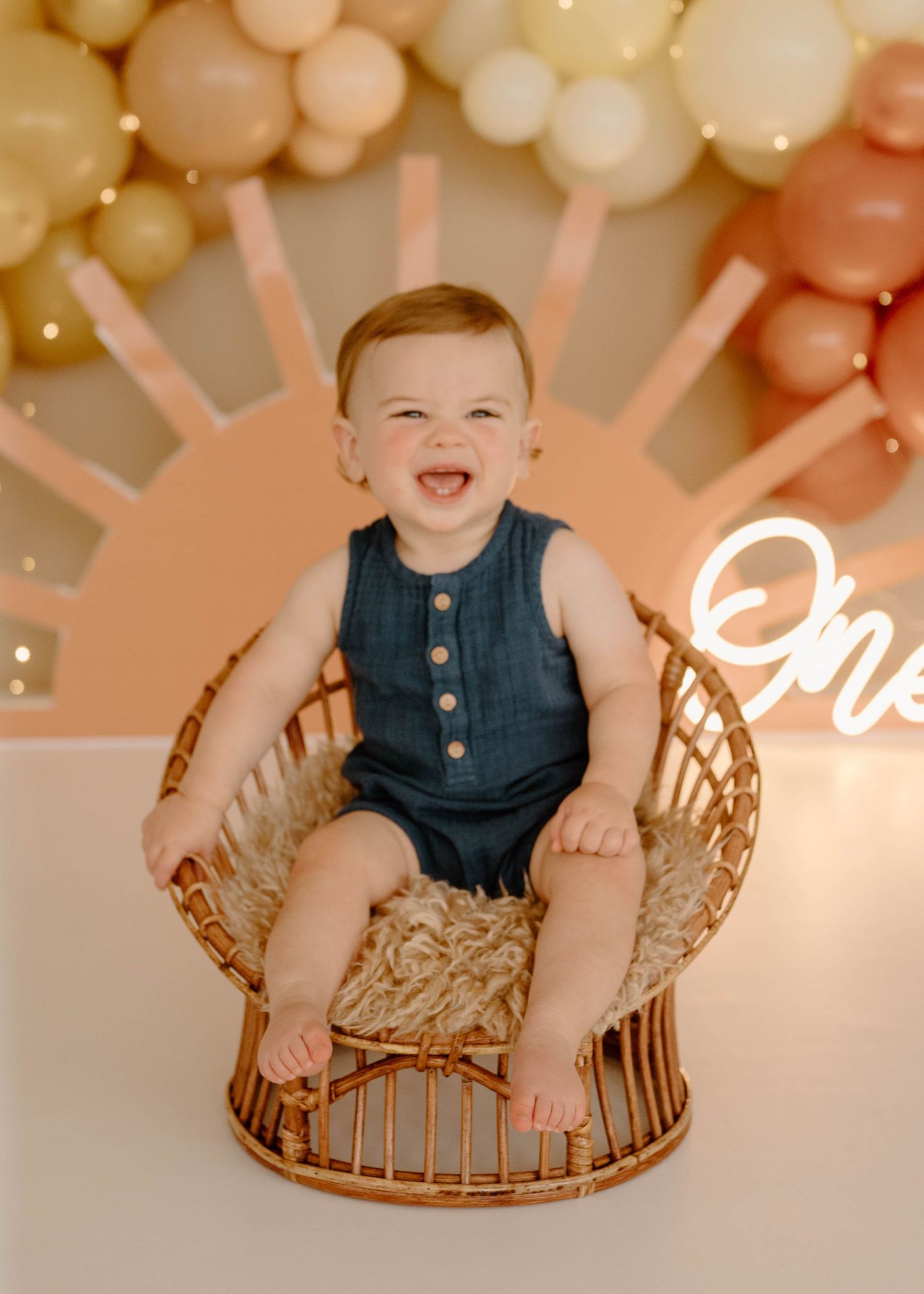 Laughing baby boy in denim romper sitting in round wicker chair with boho balloon backdrop.