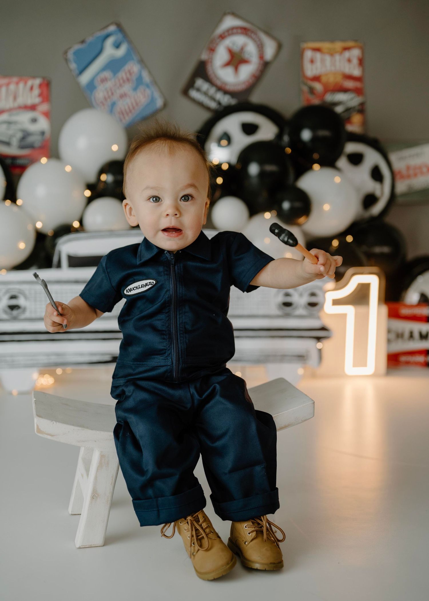 Baby boy in navy mechanic outfit sits on bench holding wrench at garage-themed first birthday session.
