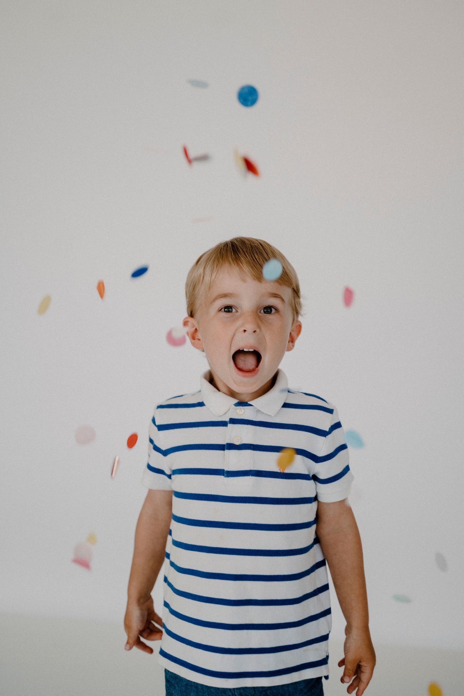 Excited young blonde boy with mouth open catching falling colorful confetti in white studio.