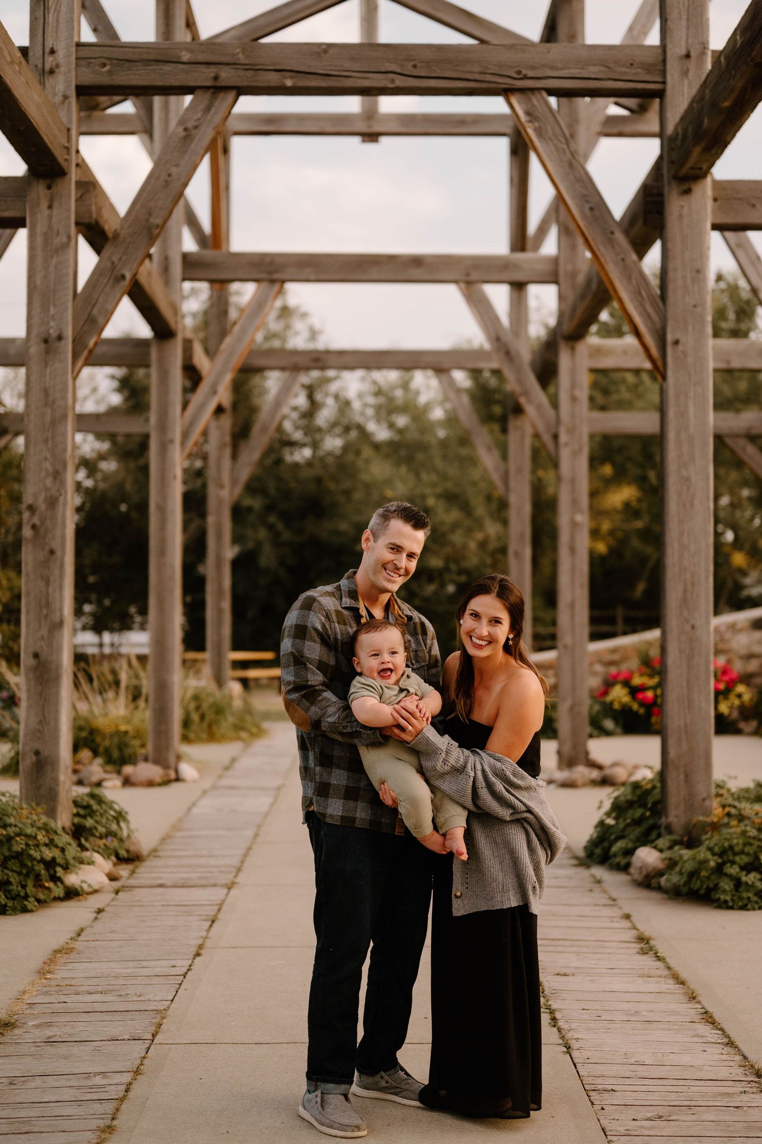 Happy family of three posing under rustic Stone Barn in Leduc surrounded by colorful garden flowers.