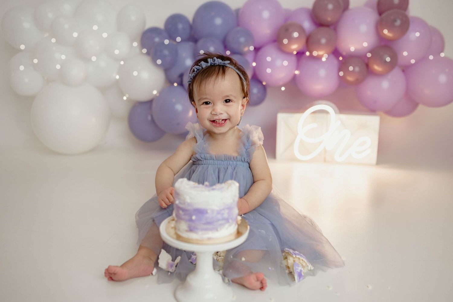 Baby girl in purple tutu smiles at first birthday cake smash with balloon garland backdrop.
