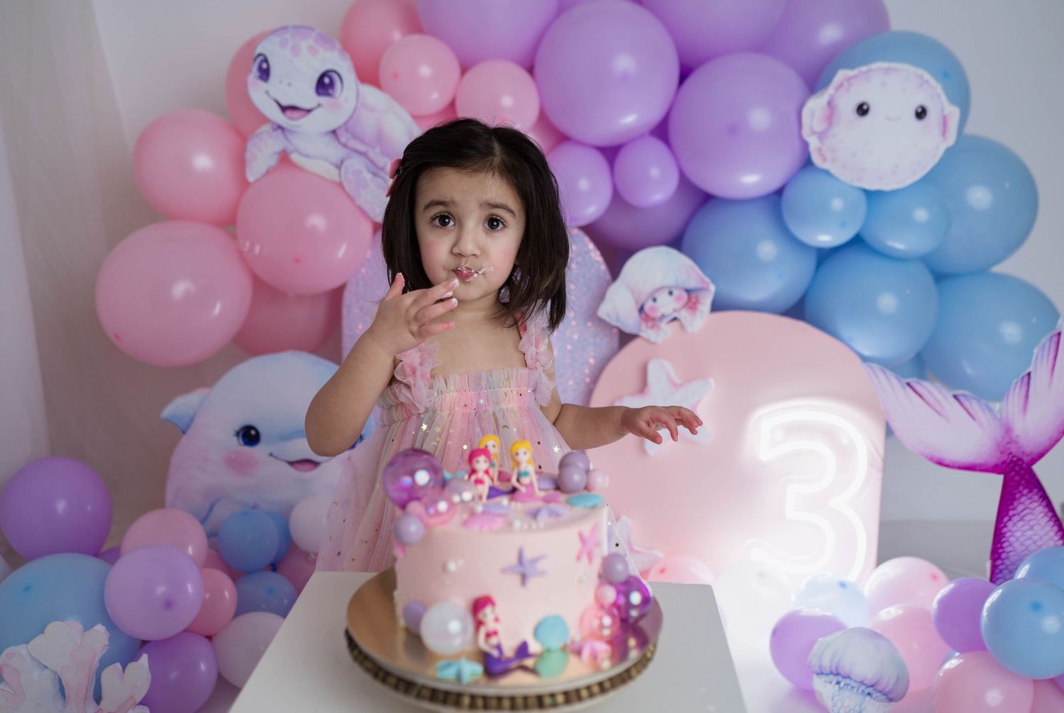 Toddler girl in pink dress eating birthday cake at mermaid themed third birthday celebration.