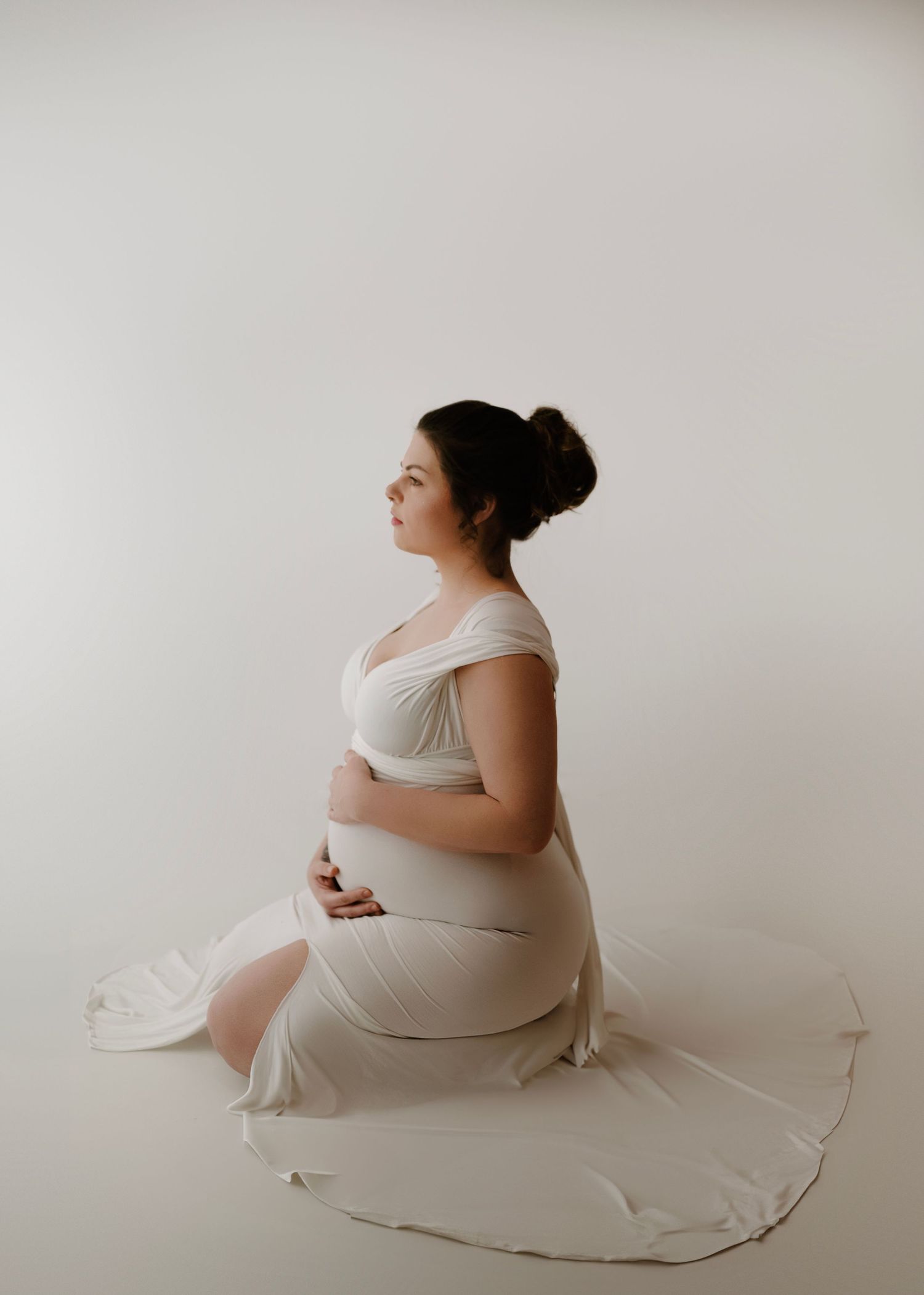 Pregnant woman in flowing white off-shoulder gown sits gracefully on floor in minimalist white studio setting.