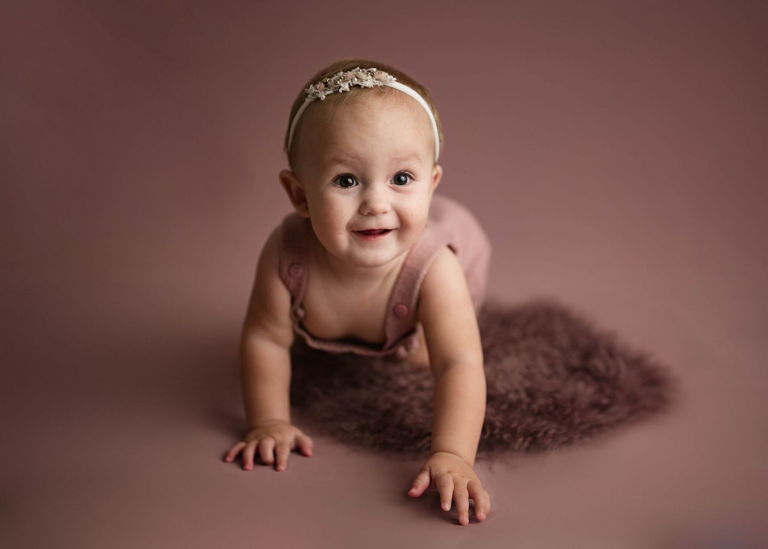Smiling baby girl in pink romper and gold headband crawling on brown fur rug in studio photo.