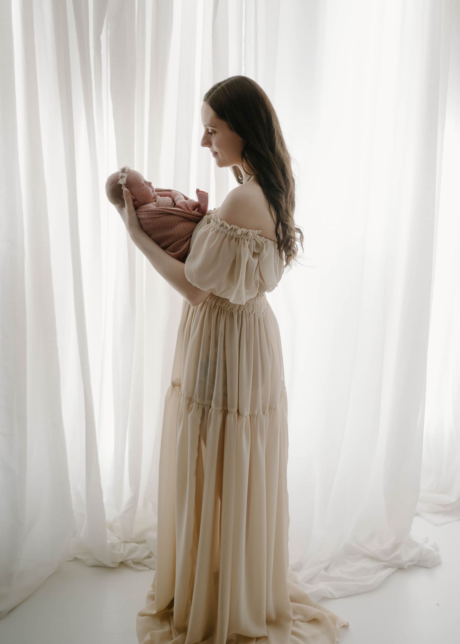 Elegant mother in cream off-shoulder gown holding newborn baby in front of white curtains for a cozy Leduc Studio session.