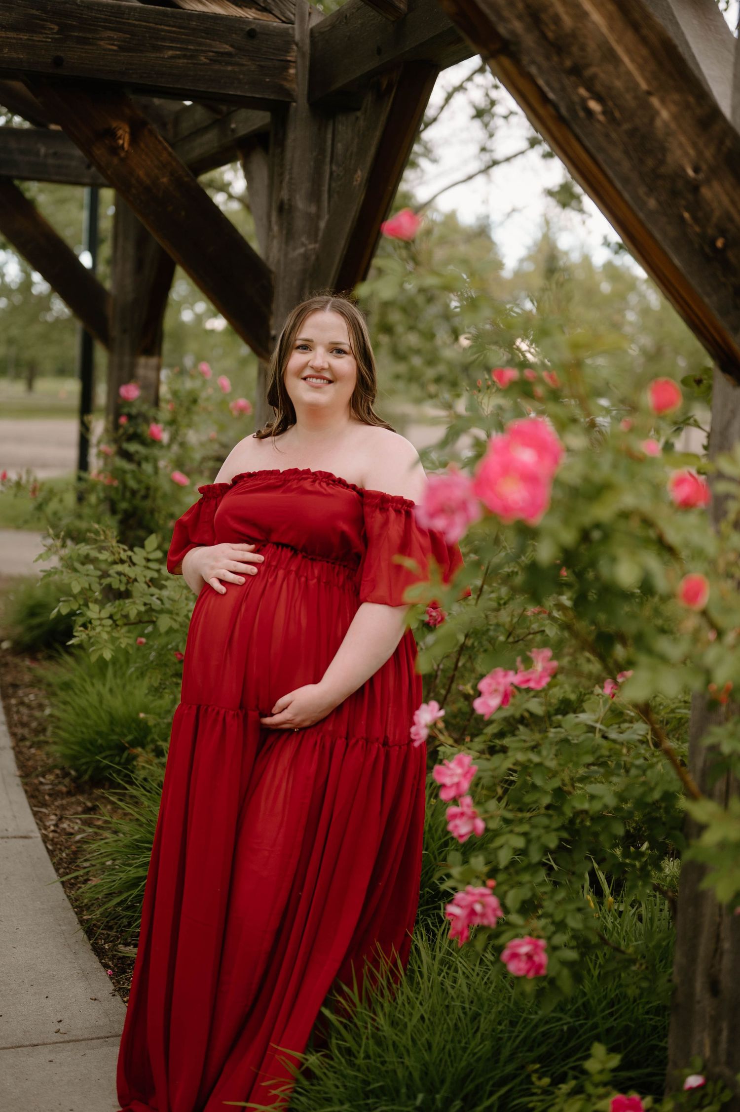 Pregnant woman in red off-shoulder gown poses among pink roses under wooden pergola for maternity photo.