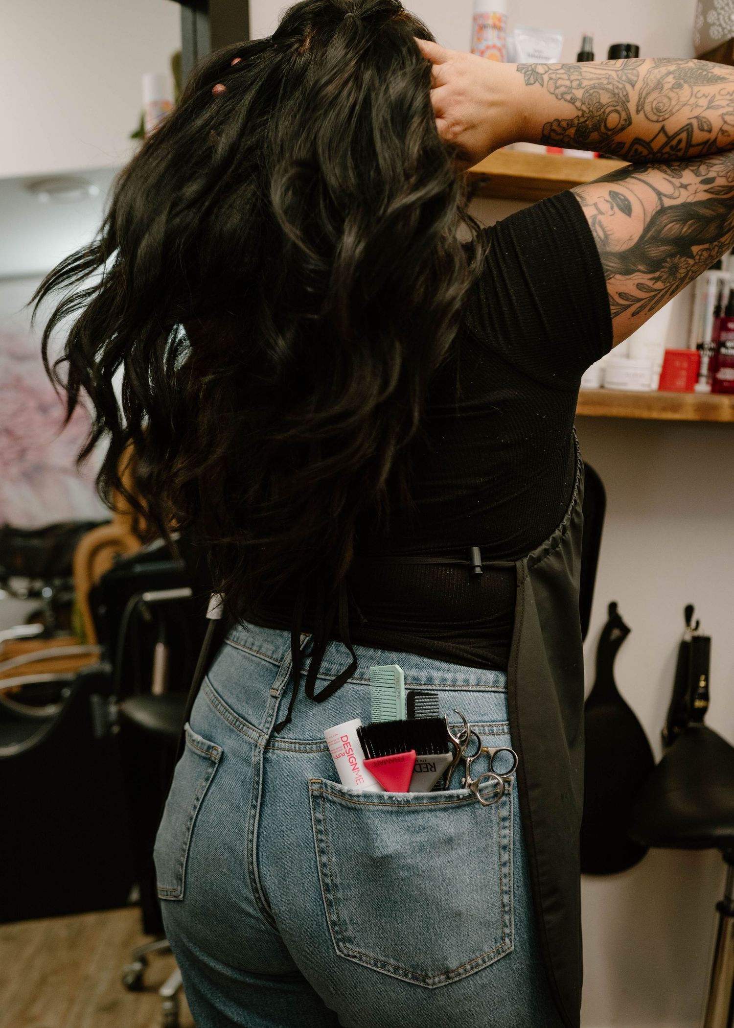 Female hairstylist with long dark wavy hair and scissors in back pocket standing in salon.