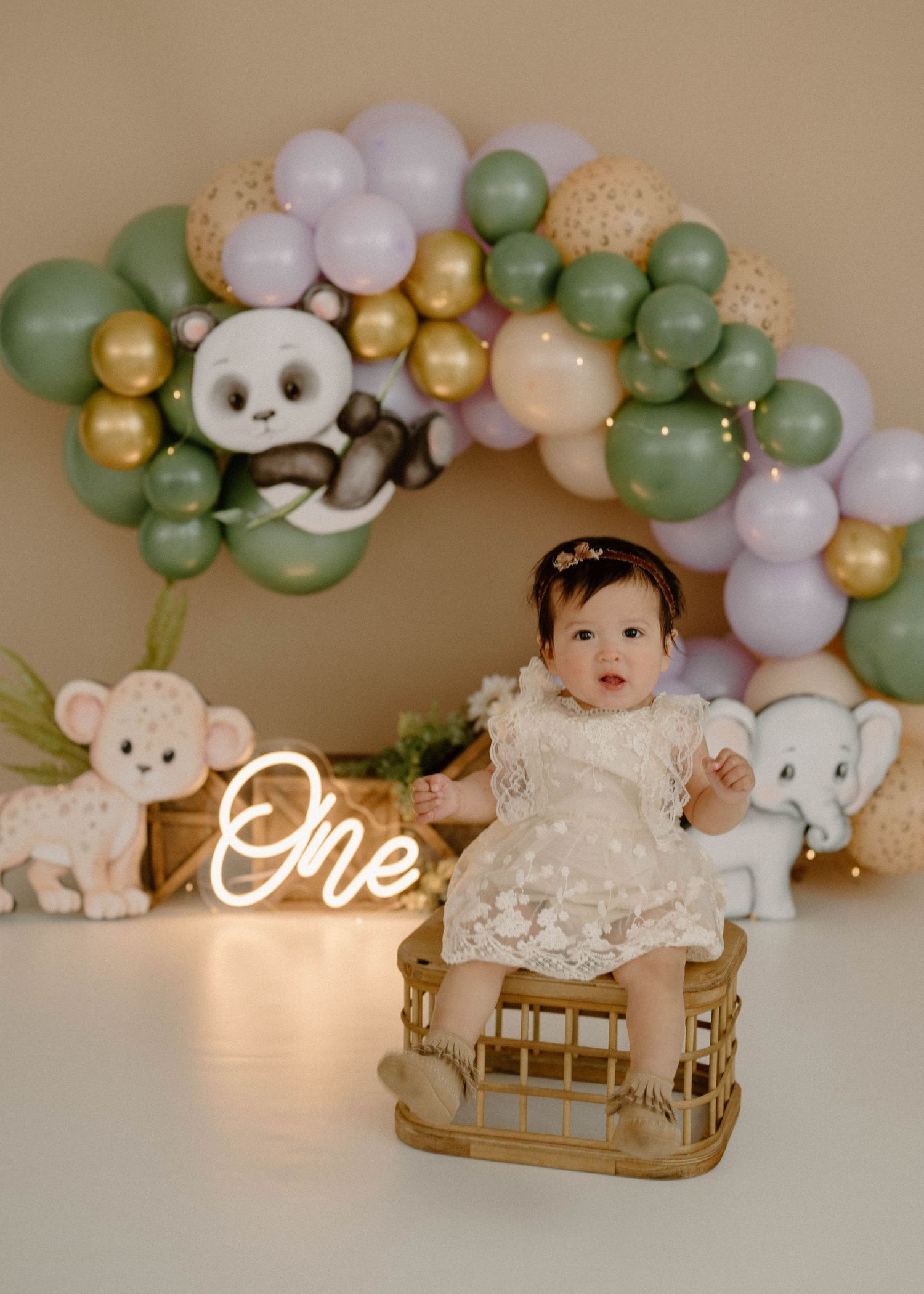 Baby girl in lace dress sits in wooden crib with panda elephant balloon arch backdrop.