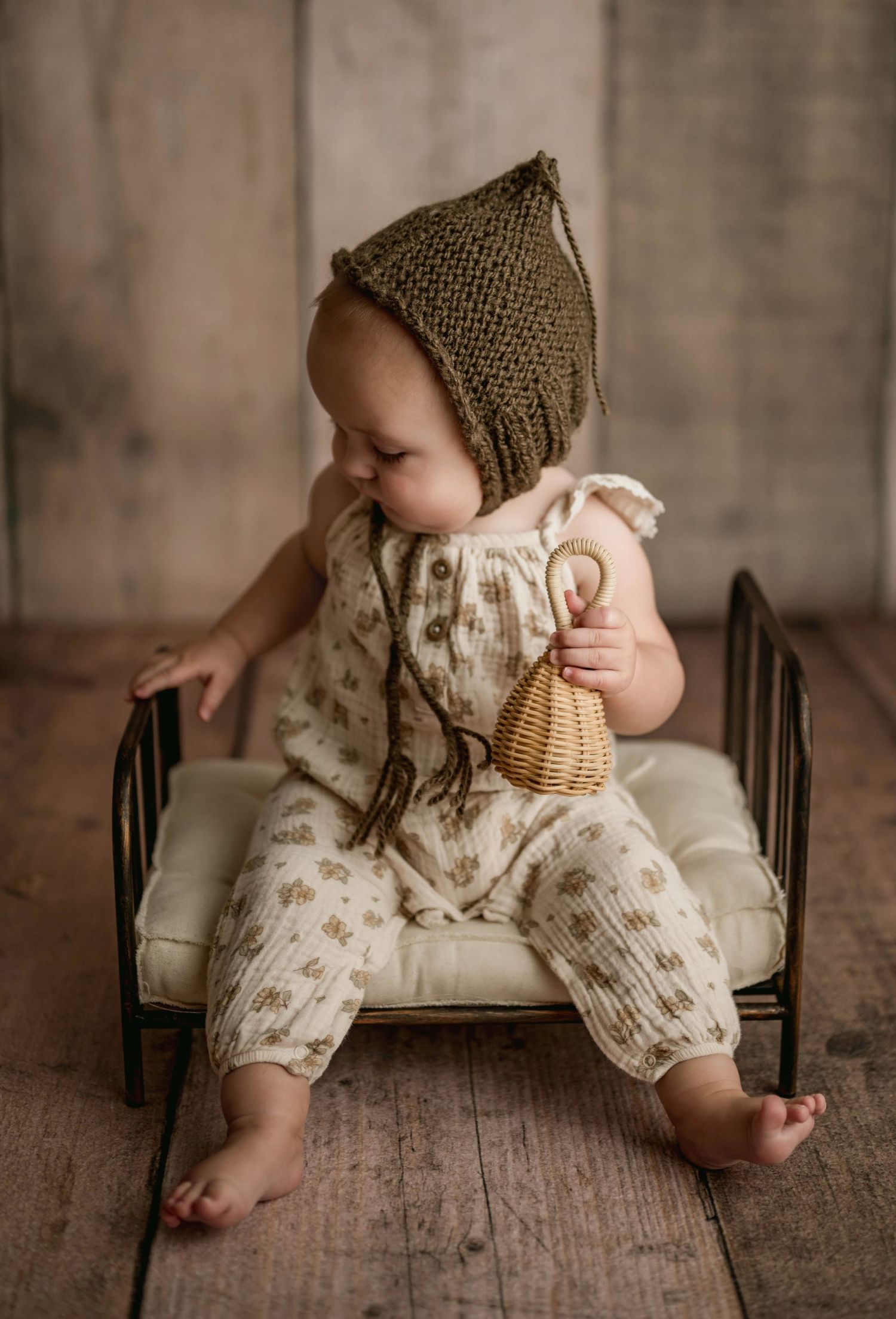 Baby in floral romper and olive knit bonnet holds wicker rattle on tiny iron bed prop.