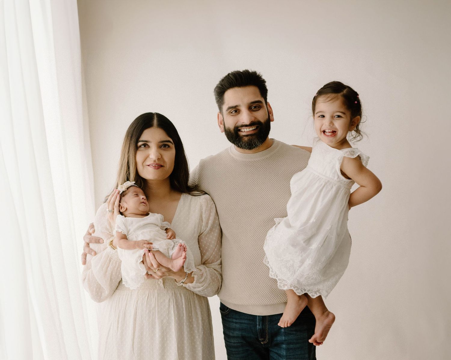 Family of four posing in studio, mother holding newborn, father holding toddler girl in white dress in Leduc Alberta studio.