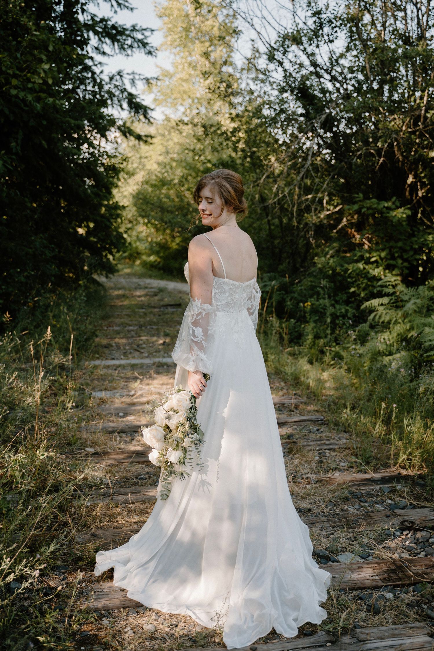 Bride in a flowing white lace wedding gown holding a bouquet, standing on a rustic path surrounded by lush greenery.