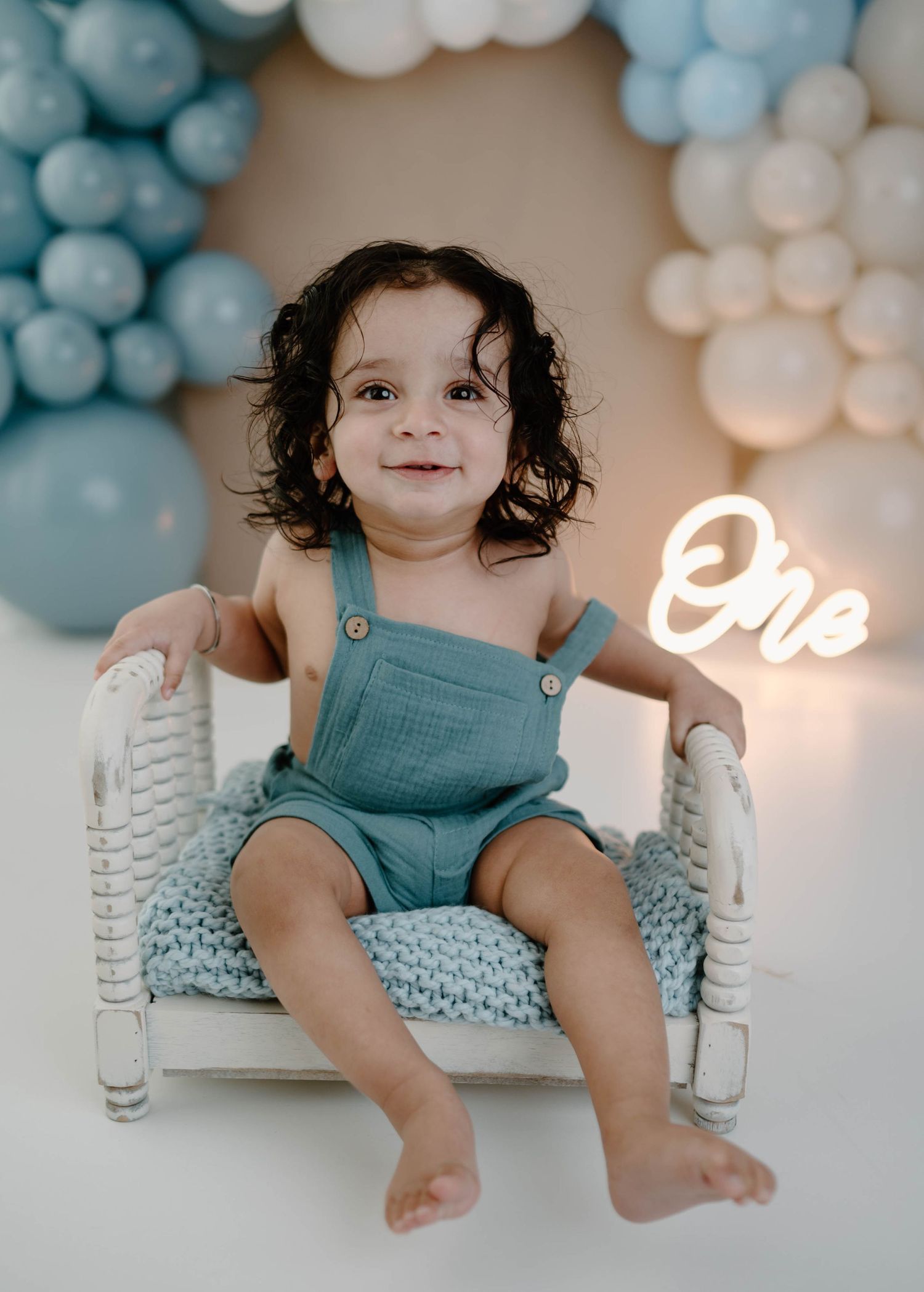 Adorable smiling baby in teal overalls sits on miniature bed with blue balloon garland and neon One sign.