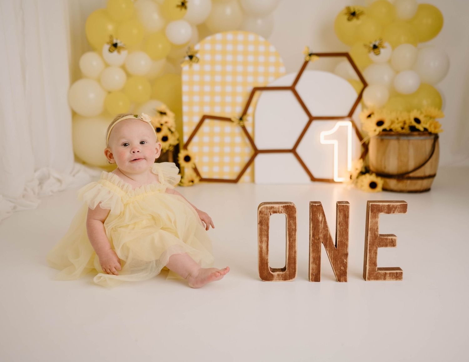 Baby girl in yellow dress sitting in front of bee-themed first birthday backdrop with ONE sign and balloons.