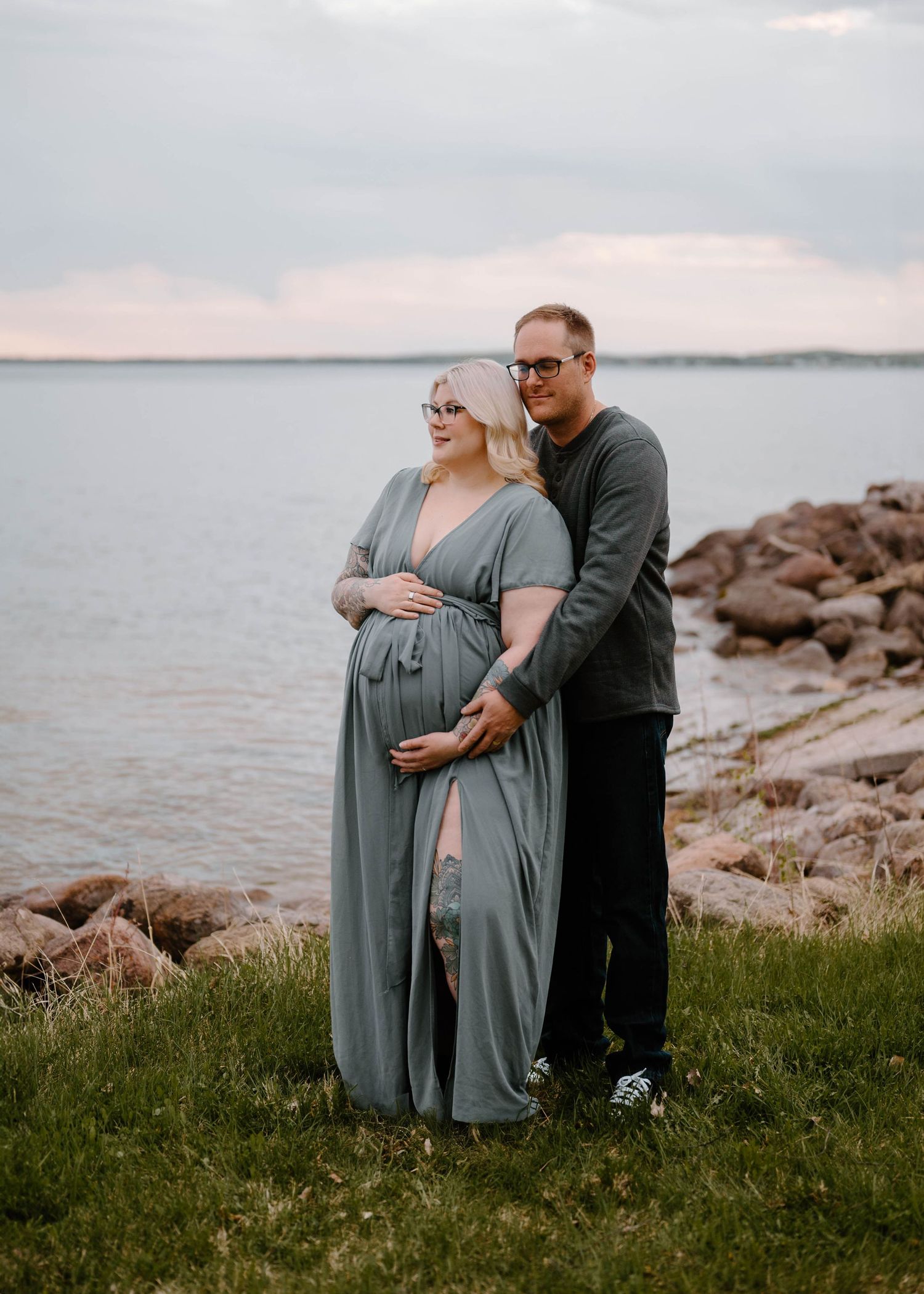 Pregnant woman in sage green maternity gown with partner by rocky lakeside at dusk.