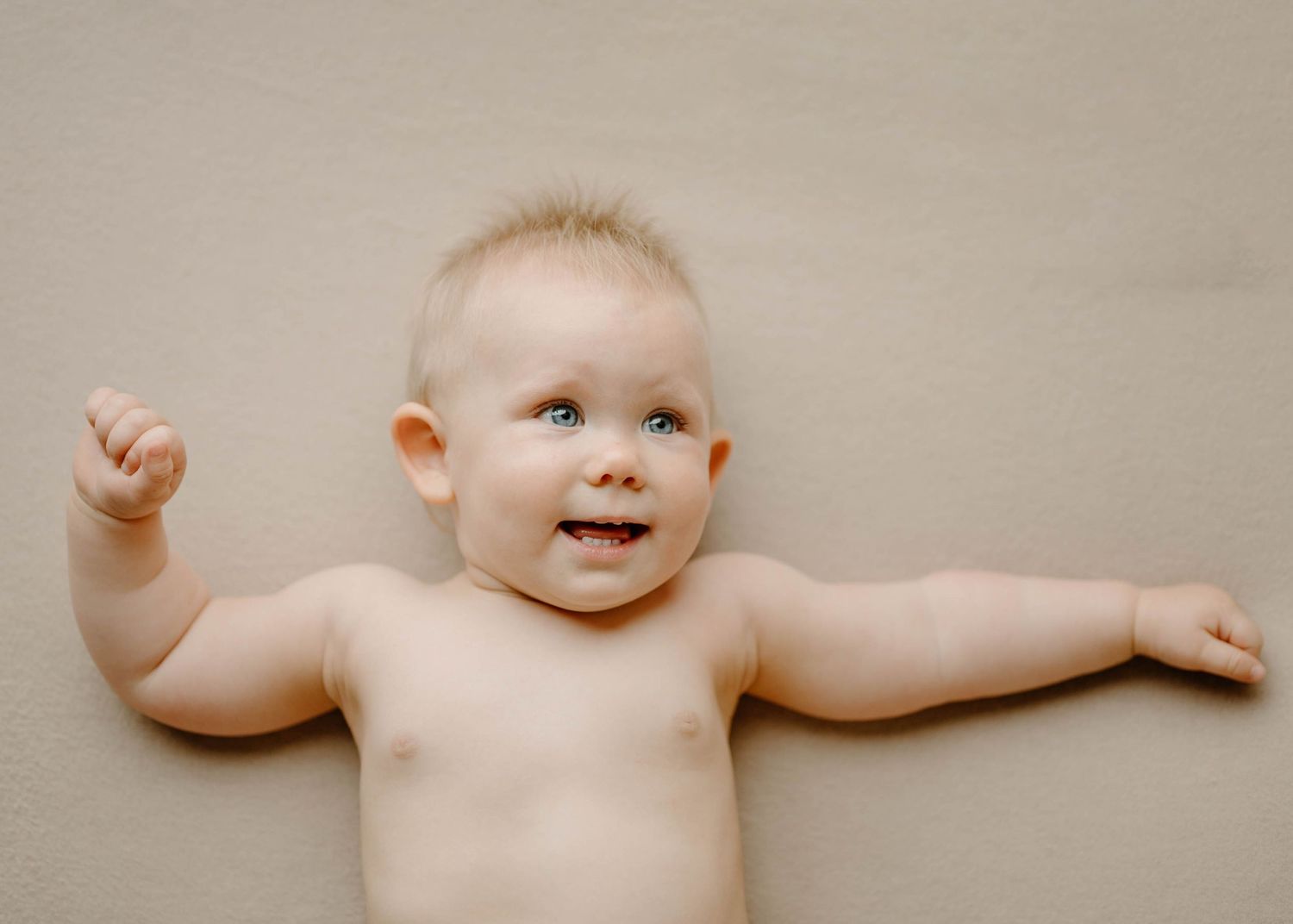 Happy baby lying on a beige backdrop with arms outstretched during a studio newborn photo session.