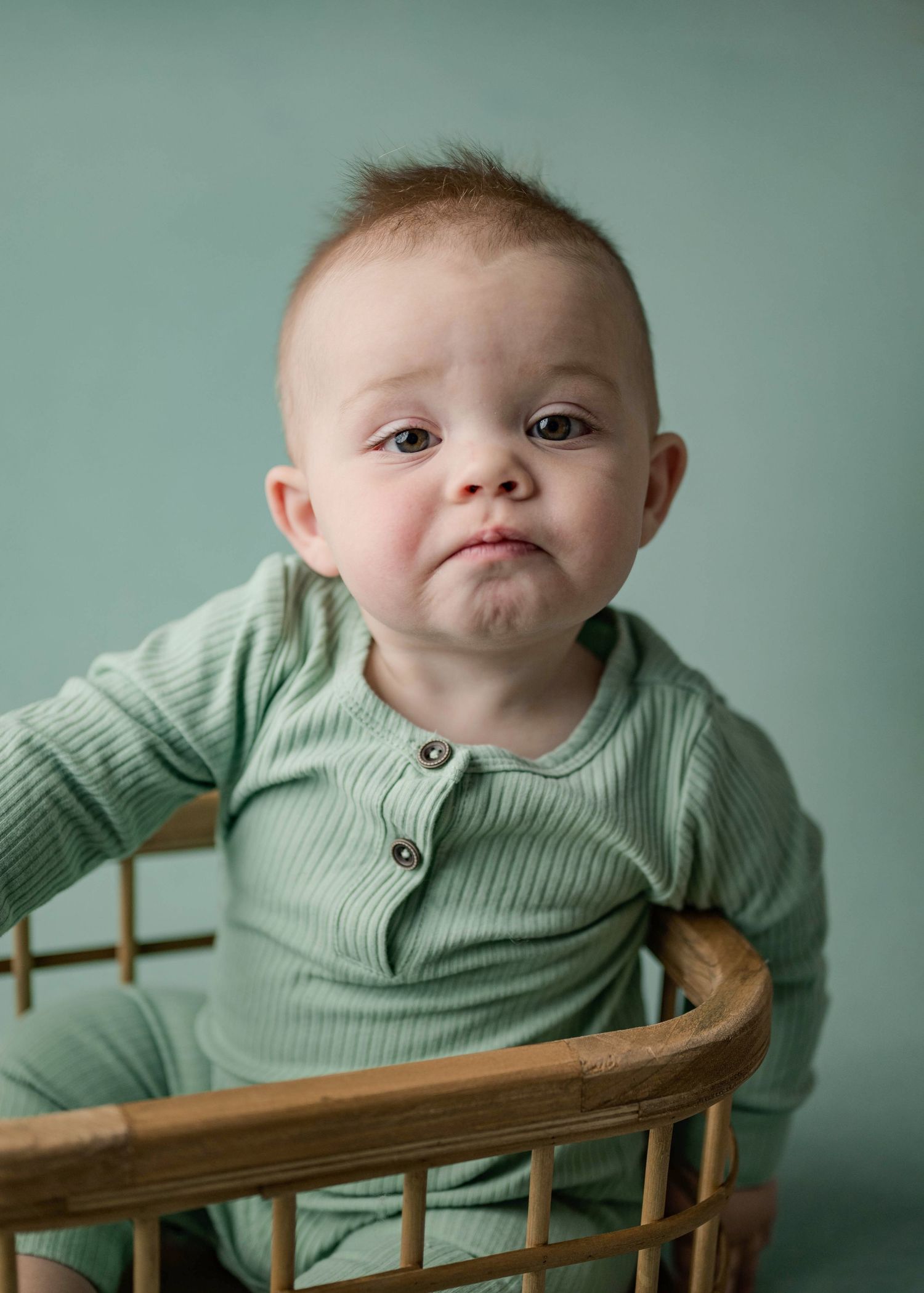 Baby boy in sage green ribbed onesie making a pouty face while sitting in a wicker basket.