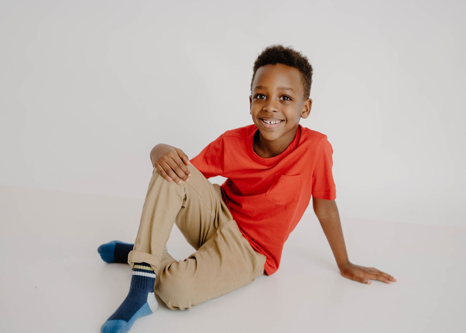 Young Black boy in a red t-shirt and khaki pants sitting casually on the floor smiling.
