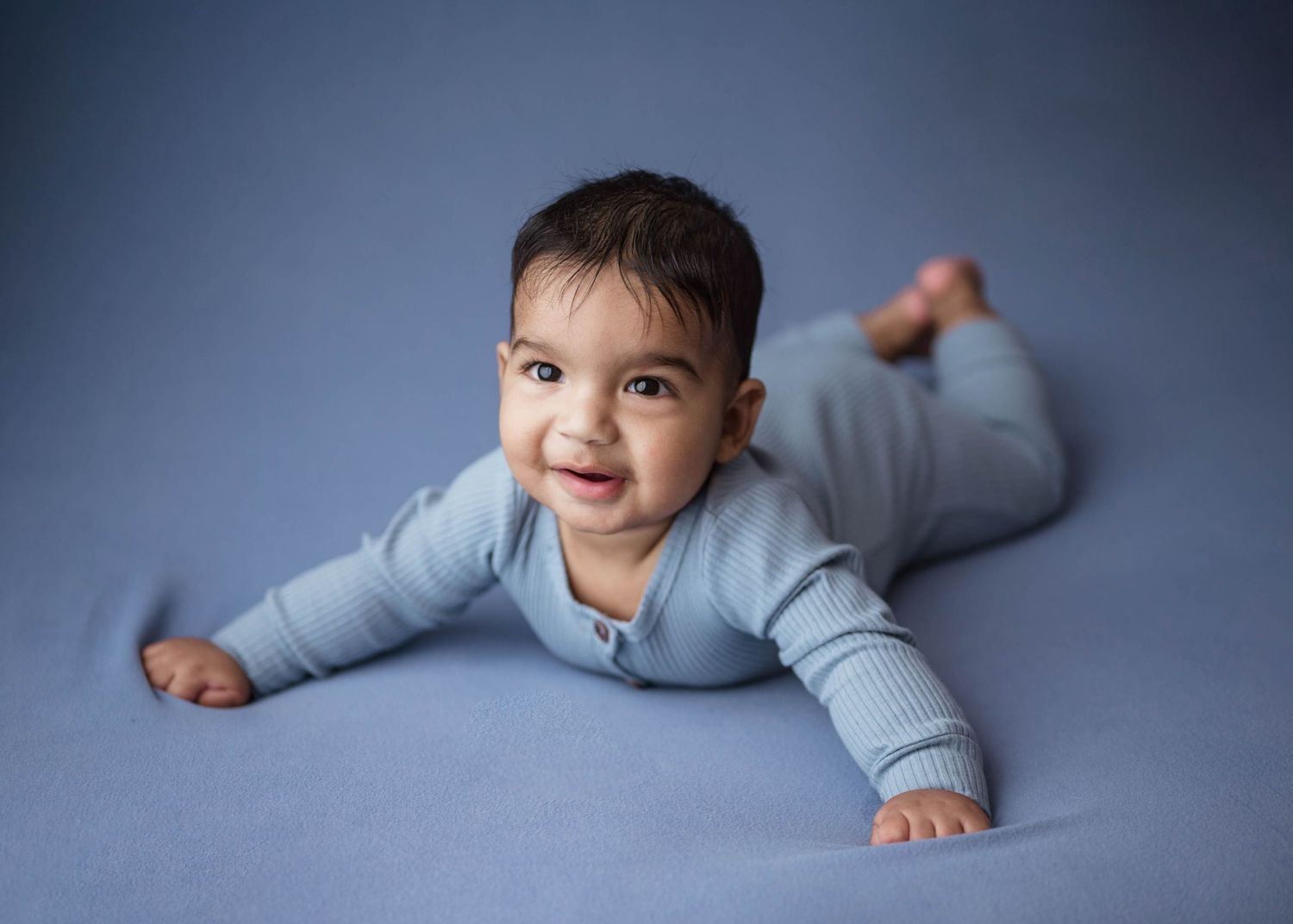 Happy baby in a blue ribbed romper doing tummy time on a soft blue blanket, smiling at the camera.