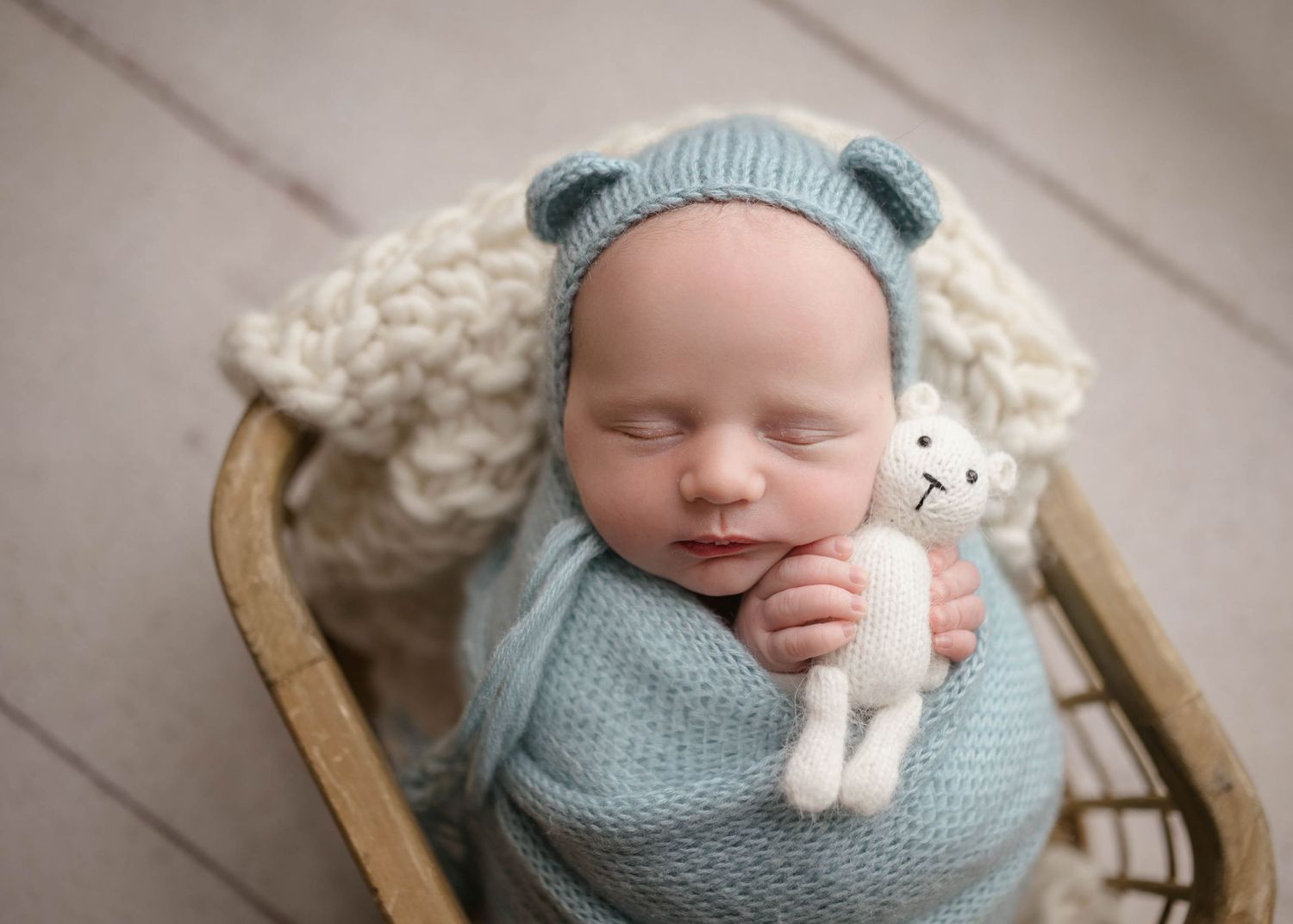 Sleeping newborn baby wrapped in blue knit blanket wearing bear bonnet holding white teddy bear in wicker basket.