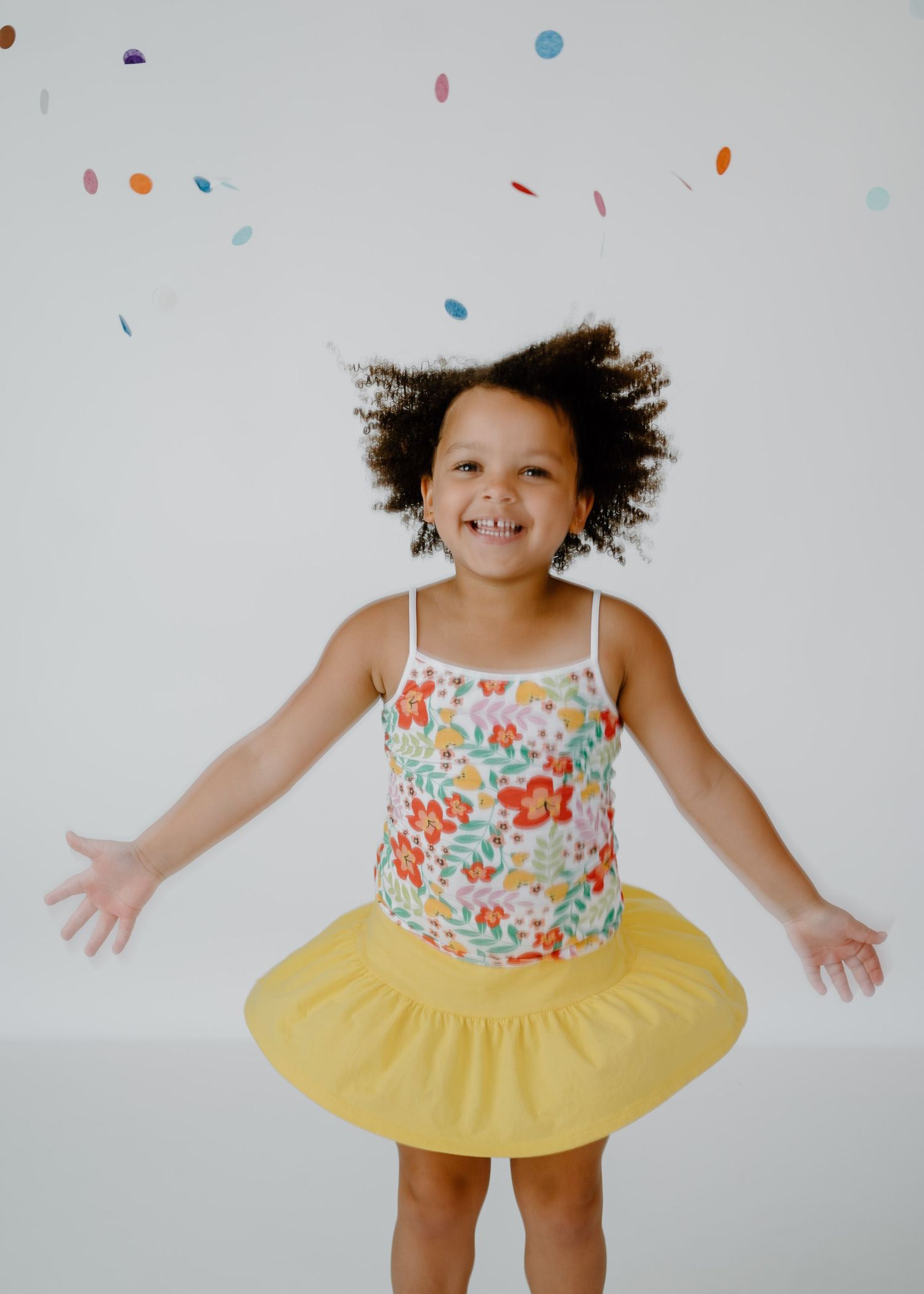 Joyful little girl in floral top and yellow skirt playing with falling colorful confetti.