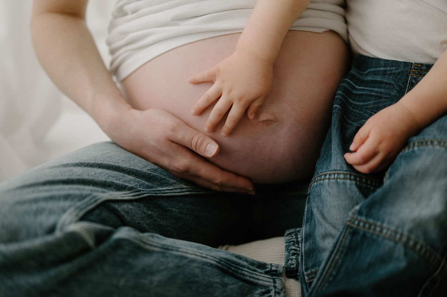 Close-up of toddler and adult hands resting on pregnant belly in jeans during maternity shoot in Leduc.